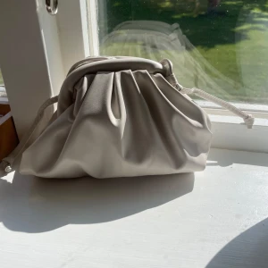 Beige dumpling bag - Säljer min beige dumpling bag med snörning upptill och mjuk, puffig form. Väskan har lång axelrem och är tillverkad i skinnimitation som ger en clean och modern vibe. Aldrig använd