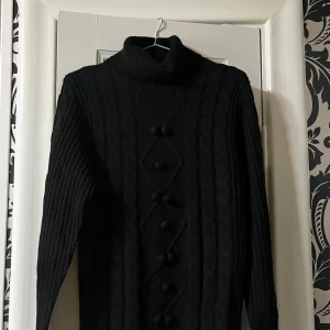 Svart polotröja med kabelstickning Boohoo - Svart polotröja från Boohoo med grov kabelstickning och dekorativa bollar längs mitt fram. Tröjan har lång ärm, ribbad polokrage och är stickad i ett mjukt material som håller dig varm. Perfekt för dig som gillar en clean och stilren look. Rak i passformen