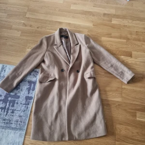 Beige kappa från Vero Moda L - Snygg beige kappa från Vero Moda i storlek L. Klassisk modell med dubbelknäppning, långa ärmar och bred krage. Jackan har två fickor framtill . Perfekt för höst och vinter när du vill vara både varm och stilren. Dock är den lite noprig därav sänkt pris.