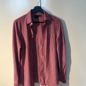 Rödvit randig skjorta Polo Ralph Lauren - Snygg röd och vit randig skjorta från Polo Ralph Lauren med klassisk krage och blå broderad logga på bröstet. Skjortan har långa ärmar, knäppning framtill och är tillverkad i mjuk bomull. Perfekt för dig som gillar stilrena och tidlösa plagg.