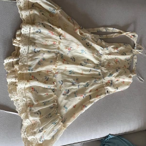 Blommig volangklänning med spetsdetaljer - Supersöt kortklänning i ljusbeige med blommigt mönster i blått och rosa. Klänningen har volanger, spetsdetaljer och knytband i ryggen. Perfekt för sommarens alla tillfällen! Materialet är mjukt och luftigt, vilket gör den extra bekväm.