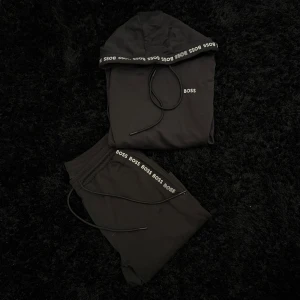 Svart dress hoodie och byxor från BOSS - Säljer en svart hoodie från BOSS med huva och dragsko. Diskret BOSS-logga på bröstet och logotypband längs huvan. Perfekt för en avslappnad och stilren look.