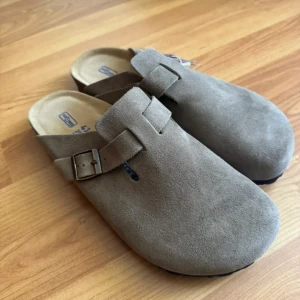 Birkenstock Boston i beige mocka - Säljer ett par Birkenstock Boston i beige mocka med justerbart spänne på sidan. Skorna har en rund tå och platt sula i kork och gummi. Perfekta för dig som gillar bekväma och stilrena tofflor med klassisk design.