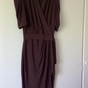Mörkbrun omlottklänning med knytband - Säljer en brun omlottklänning med knytband i midjan. Klänningen har v-ringad hals och korta ärmar med draperad effekt. Tyget faller snyggt och ger en elegant siluett. Perfekt för dig som gillar stilrena och tidlösa plagg.