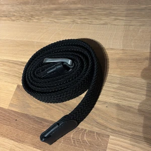 Svart flätat bälte från Jack & Jones - Säljer ett svart flätat bälte från Jack & Jones med silverfärgat spänne. Bältet har detaljer i svart skinn vid spännet och änden. Perfekt accessoar för att lyfta din outfit. Aldrig använt 