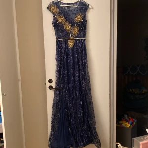 Marinblå långklänning med guldiga detaljer - Säljer en marinblå långklänning med transparent mesh upptill och vackra guldiga broderier och pärlor på framsidan. Klänningen har rund halsringning, markerad midja och glittrigt mönster över hela tyget. Perfekt för bal eller fest!