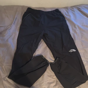 Svarta träningsbyxor från The North Face - Svarta träningsbyxor från The North Face med elastisk midja och logga på vänster ben. Byxorna har smal passform och är tillverkade i ett lätt syntetmaterial, perfekta för träning eller vardagsaktiviteter.