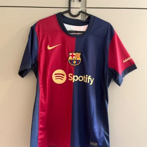 FC Barcelona matchtröja Lamine Yamal 19 - Säljer en officiell FC Barcelona matchtröja med Lamine Yamal 19 på ryggen. Tröjan är kortärmad, i rött och blått med klubbmärke, Nike-logga och sponsorer som Spotify och Ambilight tv. Materialet är lätt och andas, perfekt för fotboll.