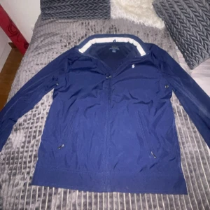 Blå vindjacka från Polo Ralph Lauren - Säljer en stilren och klassisk jacka från Polo Ralph Lauren i mörkblå färg med vit innerkrage. Jackan är i storlek barn L och i mycket fint skick, endast sparsamt använd. Den har dragkedja framtill, två sidofickor med dragkedja och den ikoniska Polo-loggan broderad på bröstet. Passar perfekt för vår och höst – lätt, bekväm och samtidigt elegant. Äkta vara, vilket framgår av etiketterna på insidan. Nypris ligger runt 1800–2000 kr. Säljer för 699 kr. Hör av er vid funderingar😁