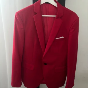 Röd kavaj med ficknäsduk - Snygg röd kavaj med klassisk passform och vit ficknäsduk. Perfekt för festliga tillfällen eller bal. Kavajen har två framfickor och en knapp framtill.