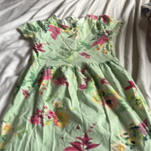 Grön blommig klänning med kort ärm - Söt ljusgrön klänning med färgglatt blommönster i rosa, gult och grönt. Klänningen har korta ärmar och en lätt utställd kjol. Perfekt för varma dagar och somriga tillfällen.
