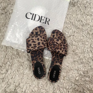 Leopardmönstrade sandaler från Cider - Säljer ett par trendiga sandaler från Cider med leopardmönster. Skorna har bred rem över foten och platt sula, perfekta för sommaren. Färgerna är bruna och svarta med djurmönster. Superlätta att bara glida i och dra ut på stan.