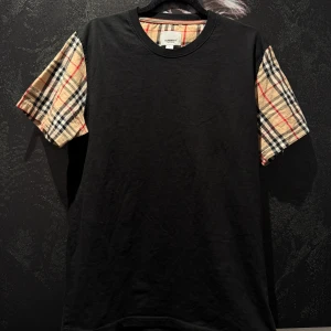 Svart t-shirt med rutiga ärmar från Burberry - Säljer en svart t-shirt från Burberry med klassiska beige-rutiga ärmar i deras ikoniska mönster. T-shirten har rund halsringning och normal passform. Perfekt för dig som vill ha en stilren look med en twist.