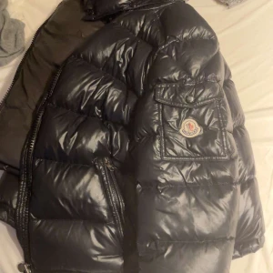 Svart pufferjacka från Moncler - Säljer en svart pufferjacka från Moncler med glansig finish och klassisk logotyp på bröstfickan. Jackan har dragkedja och flera fickor, perfekt för kyliga dagar. Stilsäker och varm modell som sticker ut.