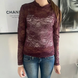 Vinröd spetsblus med lång ärm - Säljer en vinröd, transparent spetsblus med lång ärm och hög hals. Blusen har ett vackert blommigt spetsmönster och är figurnära i modellen. Perfekt att styla med linne under eller som lager på lager.