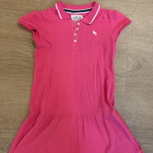 Rosa klänning  - Snygg rosa pikéklänning med klassisk krage och knappslå framtill. Klänningen har korta ärmar och liten broderad logga på bröstet. Perfekt för en avslappnad men stilren look.