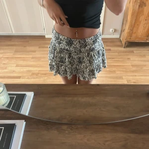 Svartvit mönstrad kortkjol - Säljer en svartvit kortkjol med grafiskt mönster. Kjolen har en lös passform och volang nertill som ger en flowy känsla. Perfekt för varma dagar och enkel att matcha med olika toppar. Storlek 134-140 men passar mig som brukar ha xs/S