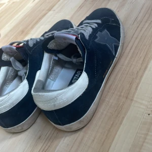 Mörkblå sneakers från Golden Goose - Säljer ett par navyblue Golden Goose sneakers. Skorna har grå snörning och vit sula. Storlek 41. Är i väldigt fint skick, en lite smutsig sula endast. Använda ett fåtal gånger