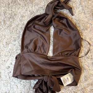 Brun omlottopp i satin - Säljer en brun omlottopp i glansigt satinmaterial med knytning både i nacken och i midjan. Toppen är ärmlös och har en djup v-ringning framtill. Perfekt för att skapa en trendig och elegant look.