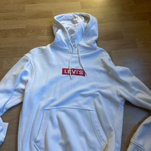 Vit hoodie från Levi's - Säljer en vit hoodie från Levi's med klassisk röd logga på bröstet. Tröjan har huva med dragsko och en stor magficka framtill. Perfekt för en avslappnad och stilren look.