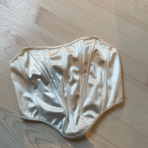 Vit satin korsett från SHEIN - Säljer en snygg vit korsett från SHEIN i glansigt satinmaterial. Toppen har markerade sömmar som ger en strukturerad look och är axelbandslös. Perfekt att styla till olika outfits!