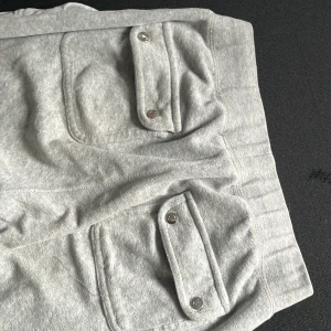 Grå Juicy Couture byxor - Säljer ett par ljusgrå mjukisbyxor från Juicy Couture med stora fickor framtill och knappar. Byxorna har resår i midjan och snörning för extra komfort. Perfekta för en avslappnad stil. 