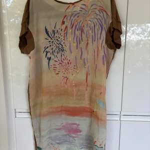 Kortklänning av Tsumori Chisato - Unik kortklänning i beige med färgglatt fyrverkerimönster framtill och enfärgad brun baksida. Klänningen har korta ärmar och en lös passform, perfekt för dig som gillar konstnärliga och lekfulla plagg. Storlek 2 (M).                                                  Tsumori Chisato is a well-known Japanese fashion designer known for her unique and playful designs that merge traditional Japanese elements with a contemporary aesthetic.