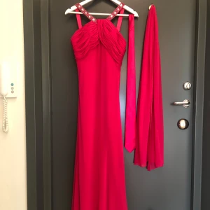 Rosa långklänning med strassdetaljer - Säljer en elegant rosa långklänning med glittriga strassdetaljer på axelbanden och vackra draperingar baktill. Klänningen har en medföljande sjal och ett snyggt fall. Perfekt för bal, bröllop eller fest. Medföljer även en slips i exakt samma färg som klänningen. 