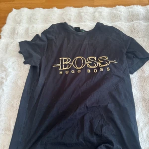 Svart t-shirt från Hugo Boss - Svart t-shirt från Hugo Boss med stor guldfärgad logga på bröstet. Klassisk rund halsringning och korta ärmar. Perfekt för en stilren och trendig look. Storlek S