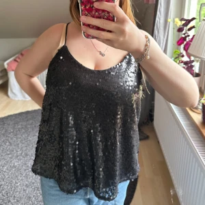 Svart paljettlinne - Säljer ett svart linne täckt med glittriga paljetter. Linnet har smala axelband och en lös passform, perfekt för att sticka ut på festen.