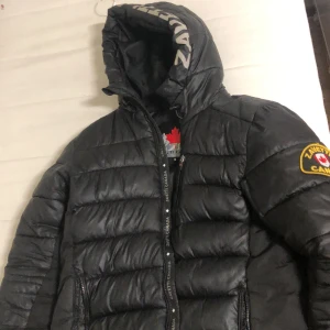 Svart pufferjacka från Zavetti Canada - Svart pufferjacka med huva från Zavetti Canada. Jackan har broderad logotyp på ärmen, dragkedja framtill och quiltad design. Perfekt för kyliga dagar med sin varma och vadderade känsla. 