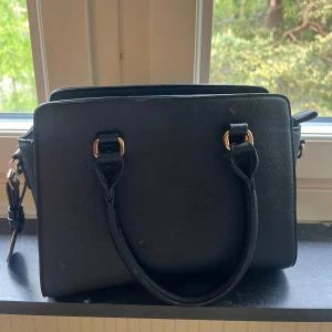 Svart handväska med gulddetaljer från Michael Kors - Snygg svart handväska från Michael Kors med guldfärgade metalldetaljer och två handtag. Väskan har en klassisk, fyrkantig form och är tillverkad i ett strukturerat syntetmaterial. Perfekt för dig som vill ha en stilren accessoar med lyxig känsla. Nypris  699kr säljer 300