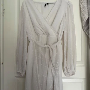 Vit omlottklänning med knytband - Säljer en vit omlottklänning med knytband i midjan och diskret prickigt mönster. Klänningen har långa ärmar med resår vid ärmslut och en v-ringad omlottdesign. Perfekt för sommarens tillfällen!