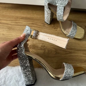 Glittriga silverklackar med bred klack - Säljer ett par riktigt snygga klackar med glitter i silver och bred fyrkantig klack. Skorna har öppen tå, justerbart spänne runt vristen och metallic guldig sula. Perfekta för att sticka ut på festen! Oanvända storlek 35