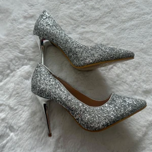 Glittriga silverfärgade pumps med klack - Säljer ett par snygga pumps i silver med glittrigt ytskikt och spetsig tå. Skorna har hög, smal klack i metallic och är perfekta för att sticka ut på festen. Klassisk pumpsmodell med platt sula och elegant form. Oanvända storlek 35 men passar mer 36