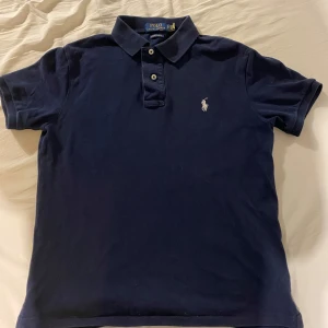 Mörkblå pikétröja från Polo Ralph Lauren - Säljer en klassisk mörkblå pikétröja från Polo Ralph Lauren med vit broderad logga på bröstet. Tröjan har korta ärmar, krage och knappar framtill. Perfekt för en stilren och avslappnad look.