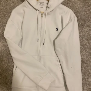 Vit hoodie från Polo Ralph Lauren - Säljer en klassisk vit kofta från Polo Ralph Lauren med dragkedja. Tröjan har två fickor framtill och den ikoniska broderade loggan i marinblått på bröstet. Är o bra skick. Enkel att matcha och riktigt snygg med rätt stil, passar samt med nästan allt samt nästan alla stilar. Säljer då jag inte använder den längre.
