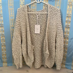 Beige blå stickad kofta från Lola & Co - Mysig beige blå stickad kofta från Lola & Co. Koftan har en grovstickad struktur och är öppen framtill utan knappar. Perfekt att slänga på sig över en topp för en avslappnad look.