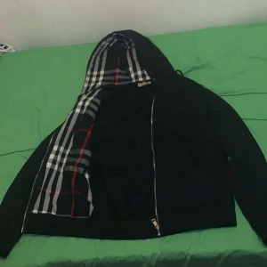 Svart hoodie från Burberry - Svart hoodie från Burberry med dragkedja och klassiskt rutigt foder i huvan. Jackan har en enkel design med fickor framtill och ikoniskt mönster i svart, vitt och rött på insidan av huvan. Tillverkad i bomull. Använd ett fåtal gånger 