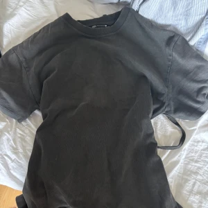 Svart omlottklänning med knytband - Säljer en mörk grå t shirt med korta ärmar och knytband i midjan. T-shirten  har en enkel och stilren design som passar perfekt till sommaren. Materialet känns mjukt och bekvämt mot huden.