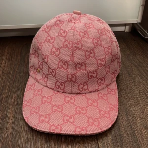 Rosa Gucci keps med GG-mönster - Snygg rosa keps från Gucci med ikoniskt GG-mönster över hela kepsen. Justerbar rem baktill i matchande rosa och liten gulddetalj på sidan. Kepsen är tillverkad i bomull och har en klassisk böjd skärm. Perfekt för dig som vill sticka ut med en lyxig accessoar.