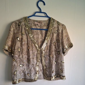 Beige bolerotopp med paljetter - Säljer en beige bolerotopp med korta ärmar och v-ringning. Toppen har ett detaljerat blommigt mönster och är dekorerad med guldiga paljetter längs kanterna och utspritt över hela plagget. Perfekt för att lyfta en outfit med lite extra glitter.