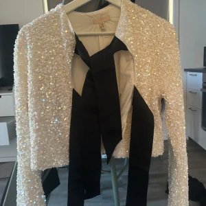 Vit paljettjacka med svart knytband - Säljer en glittrig vit jacka täckt av paljetter med lång ärm och svart knytband framtill. Jackan har en kort, croppad modell och är perfekt för att sticka ut på speciella tillfällen.