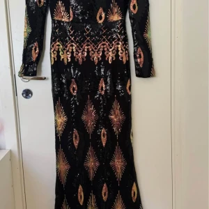 Svart långklänning med paljetter - Säljer en svart långklänning med färgglada paljettdetaljer i geometriskt mönster. Klänningen har lång ärm och figurnära passform, perfekt för festliga tillfällen. Fyrkantig ringning och elegant design som verkligen sticker ut.