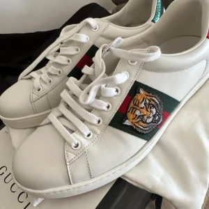 Gucci ace skor - Säljer ett par vita Gucci skor med klassiska gröna och röda ränder på sidorna samt en broderad tiger. Skorna har snörning och metallic-detaljer i rött och grönt på hälen. Perfekta för dig som vill sticka ut med lyxiga detaljer. Skorna är knappt använda endast fåtal gånger. Allt OG ingår förutom kvitto däremot finns det vestiaire collective äkthet tag alltså verifiering av experter att de är autentiska!  Skorna är självfallet äkta!