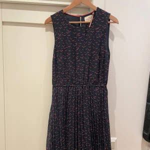 Mörkblå klänning med mönster från Holly & Whyte - Snygg mörkblå ärmlös klänning från Holly & Whyte med små rödvita mönster. Klänningen har rund halsringning och markerad midja, perfekt för en stilren look. Lätt och luftig modell som passar till många tillfällen.