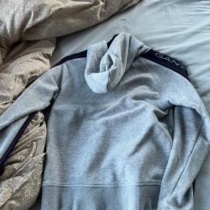 Grå hoodie från Gant - Säljer en ljusgrå hoodie från Gant med huva och magficka. Tröjan har mörkblå ränder med Gant-logga längs ärmarna och är tillverkad i mjukt material. Perfekt för en avslappnad stil.
