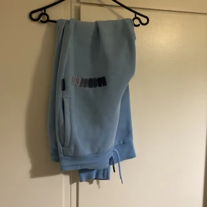 Ljusblå mjukisbyxoroch hoodie från Hoodrich - Säljer ett par ljusblå mjukisbyxor från Hoodrich med broderad logga på ena benet. Byxorna har resår i midjan med dragsko och sidofickor. Perfekta för en avslappnad stil.