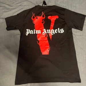 Palm Angels x VLONE - Svart t-shirt med stor vit och röd VLONE-logga på framsidan och Palm Angels-tryck på ryggen. T-shirten har rund hals och korta ärmar. Perfekt för dig som gillar streetwear och vill sticka ut med en unik design.