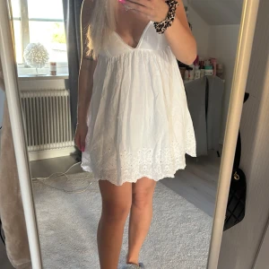 Vit kortklänning med broderade detaljer - Söt vit kortklänning med broderade blommor nertill och v-ringad urringning. Klänningen är ärmlös och har en luftig passform, perfekt för varma dagar. Tillverkad i bomull för en lätt och bekväm känsla.